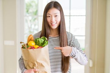 Beautiful Asian woman holding paper bag of fresh groceries very happy pointing with hand and finger