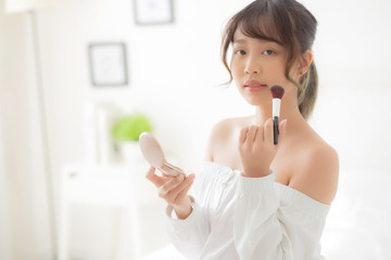 Beauty portrait young asian woman smiling with face looking mirror applying makeup with brush cheek in the bedroom, beautiful girl holding blusher facial, skin care and cosmetic concept.
