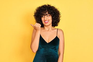 Young arab woman with curly hair wearing elegant dress over isolated yellow background smiling with happy face looking and pointing to the side with thumb up.