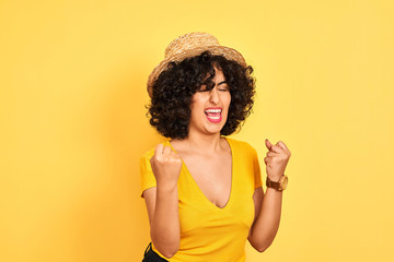 Young arab woman with curly hair wearing t-shirt and hat over isolated yellow background very happy and excited doing winner gesture with arms raised, smiling and screaming for success. 