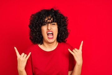 Young arab woman with curly hair wearing casual t-shirt over isolated red background shouting with crazy expression doing rock symbol with hands up. Music star. Heavy concept.