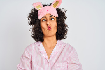 Arab woman with curly hair wearing pajama and sleep mask over isolated white background making fish face with lips, crazy and comical gesture. Funny expression.