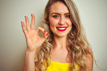 Young beautiful woman wearing yellow t-shirt standing over white isolated background doing ok sign with fingers, excellent symbol