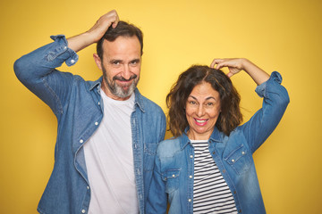 Beautiful middle age couple together wearing denim shirt over isolated yellow background confuse and wonder about question. Uncertain with doubt, thinking with hand on head. Pensive concept.