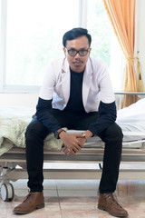 Portrait of Asian young male doctor with white lab coat sitting on indoors hospital clinic bed looking at camera