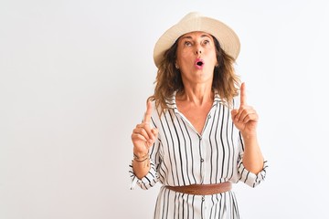 Middle age businesswoman wearing striped dress and hat over isolated white background amazed and surprised looking up and pointing with fingers and raised arms.