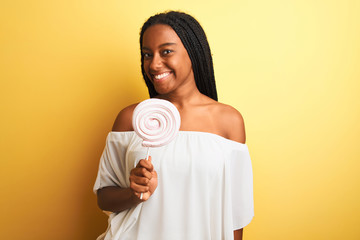 Young african american woman eating candy standing over isolated yellow background with a happy face standing and smiling with a confident smile showing teeth