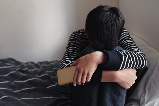 Young Asian preteen teenager boy hugging his knee in his bedroom with smartphone, FOMO, Cyber bullying in kid, depressed child mental illness health