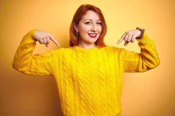 Beautiful redhead woman wearing winter sweater standing over isolated yellow background looking...