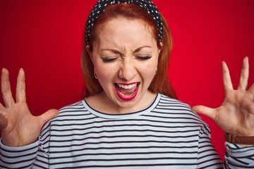Beautiful redhead woman wearing striped navy t-shirt standing over isolated red background celebrating mad and crazy for success with arms raised and closed eyes screaming excited. Winner concept