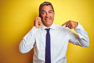 Middle age businessman talking on the smartphone over isolated yellow background with surprise face pointing finger to himself