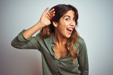 Young beautiful woman wearing green shirt standing over grey isolated background smiling with hand over ear listening an hearing to rumor or gossip. Deafness concept.