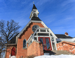 Orange brick church in rural midwest. Red doors, white trim & steeple topped with a cross. Large tree without leaves and blue sky.