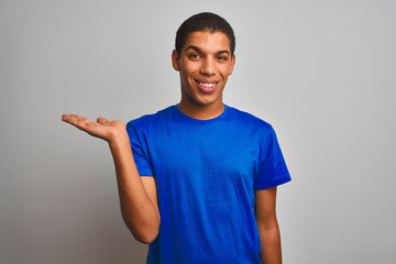Fototapeta premium Young handsome arab man wearing blue t-shirt standing over isolated white background smiling cheerful presenting and pointing with palm of hand looking at the camera.