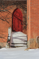 Red door in brick building, snow covered stairs, with dinstinct shadows from bare tree branches/