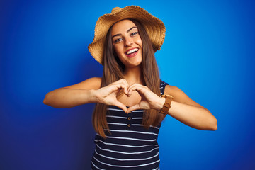 Young beautiful woman wearing striped t-shirt and summer hat over isolated blue background smiling in love showing heart symbol and shape with hands. Romantic concept.