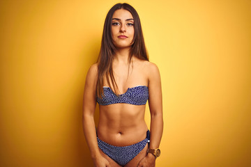 Young beautiful woman on vacation wearing bikini standing over isolated yellow background Relaxed with serious expression on face. Simple and natural looking at the camera.