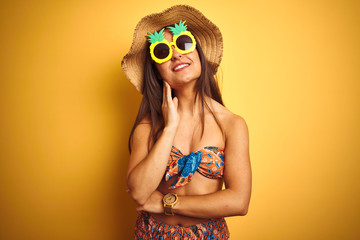 Woman on vacation wearing bikini and pineapple sunglasses over isolated yellow background with hand on chin thinking about question, pensive expression. Smiling with thoughtful face. Doubt concept.