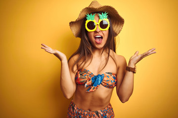 Woman on vacation wearing bikini and pineapple sunglasses over isolated yellow background celebrating crazy and amazed for success with arms raised and open eyes screaming excited. Winner concept