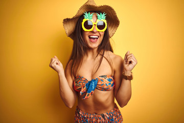 Fototapeta premium Woman on vacation wearing bikini and pineapple sunglasses over isolated yellow background celebrating surprised and amazed for success with arms raised and open eyes. Winner concept.