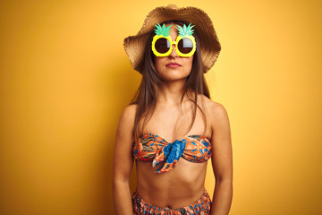 Woman on vacation wearing bikini and pineapple sunglasses over isolated yellow background with serious expression on face. Simple and natural looking at the camera.
