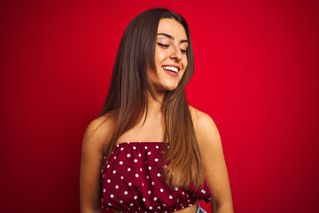 Young beautiful woman wearing casual t-shirt standing over isolated red background looking away to side with smile on face, natural expression. Laughing confident.