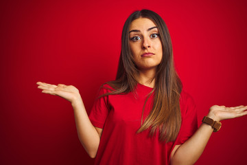 Young beautiful woman wearing t-shirt standing over isolated red background clueless and confused...