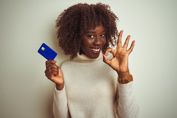 Young african afro woman holding credit card standing over isolated white background doing ok sign...