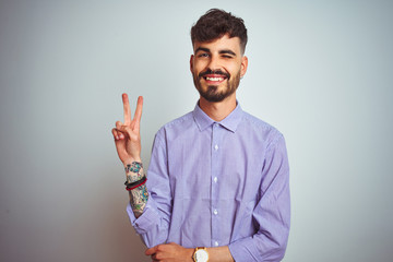 Young man with tattoo wearing purple shirt standing over isolated white background smiling with happy face winking at the camera doing victory sign. Number two.