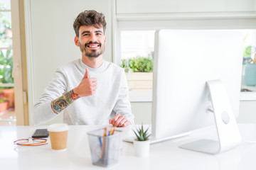 Young man working using computer doing happy thumbs up gesture with hand. Approving expression looking at the camera with showing success.