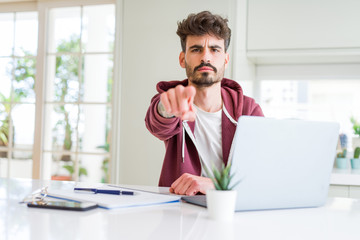 Young student man using computer laptop and notebook pointing with finger to the camera and to you, hand sign, positive and confident gesture from the front