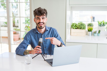 Young man recording podcast using microphone and laptop very happy pointing with hand and finger