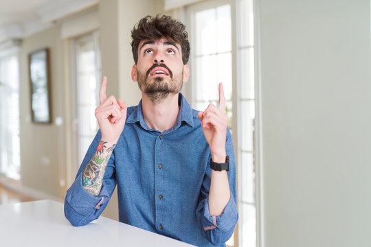 Young man wearing casual shirt sitting on white table amazed and surprised looking up and pointing with fingers and raised arms.