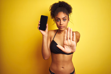 Fototapeta premium African american woman wearing bikini showing smartphone over isolated yellow background with open hand doing stop sign with serious and confident expression, defense gesture