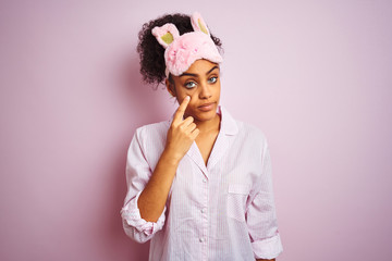Young african american woman wearing pajama and mask over isolated pink background Pointing to the eye watching you gesture, suspicious expression
