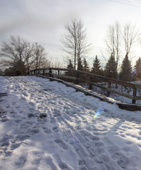 Mid winter in the mid west. Snow covered ground, wooden fence, pine trees, tall trees with no leaves and a setting sun.