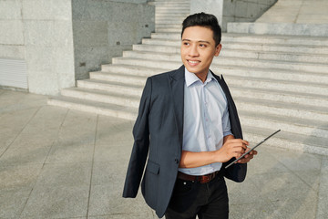 Portrait of cheerful young Asian businessman working on tablet computer when standing outdoors