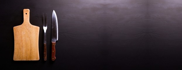 Banner para website com tábua de corte em madeira natural, faca de carne e garfo antigos sobre a...