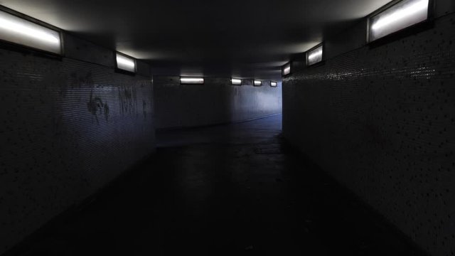 Handheld footage of person walking scared through dim lit underpass towards light at end of tunnel with person coming towards