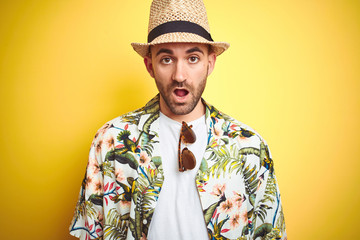 Young man on vacation wearing hawaiian flowers shirt and summer hat over yellow background afraid...