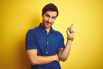 Young handsome man with tattoo wearing casual shirt standing over isolated yellow background with a big smile on face, pointing with hand and finger to the side looking at the camera.