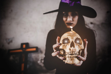 Halloween concept - Witch woman holding a ghost skull, Mysterious demon girl with red lips. Vampire Witch lady with blood in mouth on black background.