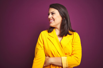 Young beautiful brunette woman wearing elegant yellow jacket over purple isolated background looking away to side with smile on face, natural expression. Laughing confident.