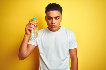 Young brazilian man holding bottle of water standing over isolated yellow background scared in shock with a surprise face, afraid and excited with fear expression