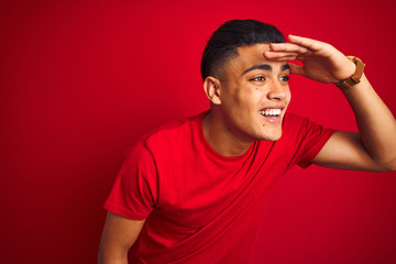 Young brazilian man wearing t-shirt standing over isolated red background very happy and smiling looking far away with hand over head. Searching concept.