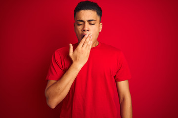 Young brazilian man wearing t-shirt standing over isolated red background bored yawning tired covering mouth with hand. Restless and sleepiness.