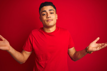 Young brazilian man wearing t-shirt standing over isolated red background clueless and confused expression with arms and hands raised. Doubt concept.