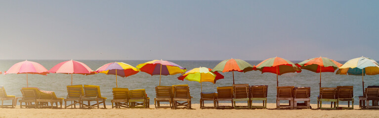 Horisontal banner - row of empty wooden chaise lounges with colorful umbrellas on the beach in GOA, India, toned with sunlight. Sea summer holiday background. - obrazy, fototapety, plakaty