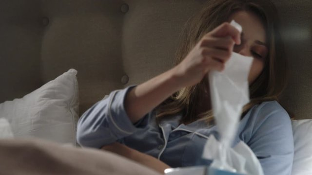 A sick, suffering woman in bed with a virus blows her nose using a tissue then takes a sip of hot tea.