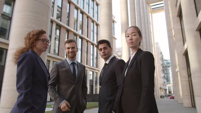 Tilt down of two diverse businesswomen talking to two elegant businessmen standing nearby in downtown and then looking at camera